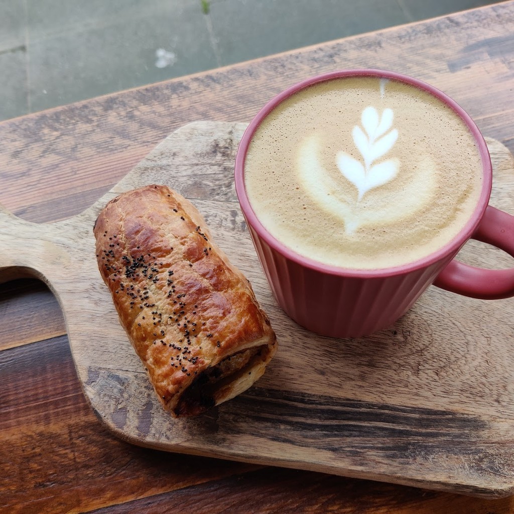 A flat white in a pink ribbed mug beside a sausage roll on a wooden board