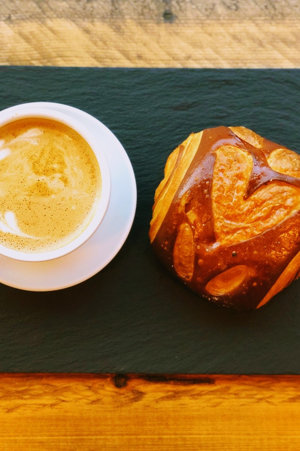 A cappuccino with latte art beside a pretzel-style bread roll on a slate board